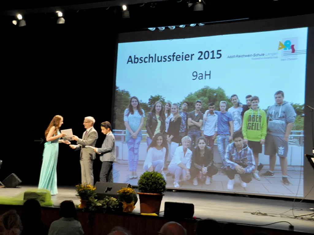 Eine Übergabe auf der Bühne vor dem projizierten Klassenfoto.